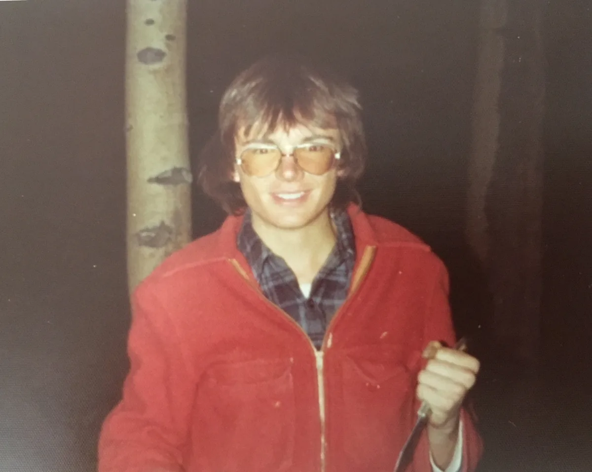 Vintage flash photograph of a smiling young man with shaggy hair and amber-tinted aviator glasses, wearing a red jacket and holding a knife, outdoors at night